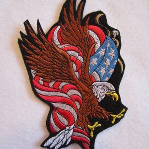 May include: Embroidered patch featuring a brown eagle with outstretched wings, holding an American flag in its talons. The eagle is surrounded by red, white, and blue swirling designs.