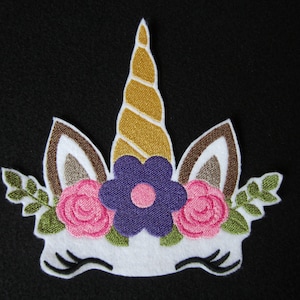 May include: Embroidered unicorn head patch with a gold horn, purple flower, and pink flowers. The unicorn has brown ears and white fur.