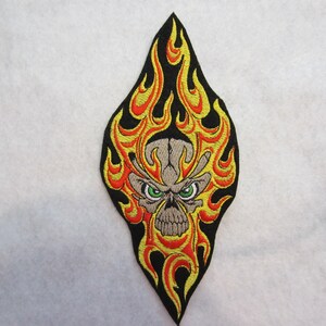 May include: Embroidered patch featuring a skull with green eyes and flames in shades of yellow, orange, and red. The skull is gray, and the flames are outlined in black. The patch has a teardrop shape.