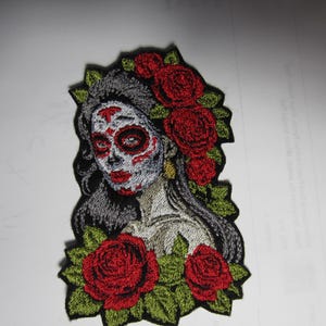 May include: Embroidered patch featuring a stylized depiction of a woman with a skull-like face, adorned with red roses and green leaves. The woman has long gray hair and is wearing makeup. The patch is likely for clothing or accessories.