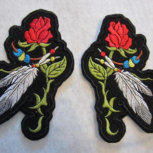 May include: Two identical embroidered patches on a black background, each featuring a red rose with green leaves, two white feathers, and a string of blue, yellow, and red beads. The design is outlined in black thread with a scalloped edge.