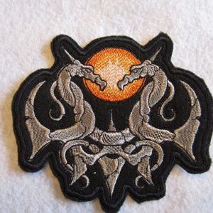 May include: Embroidered patch featuring two silver dragons facing each other with their tails intertwined. The dragons are silhouetted against a black background with an orange sun in the center.