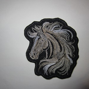 May include: Embroidered patch featuring a horse head profile with a flowing mane. The horse is depicted in shades of brown and silver against a black background. The patch has a black, irregular border, suitable for sewing or ironing onto fabric.