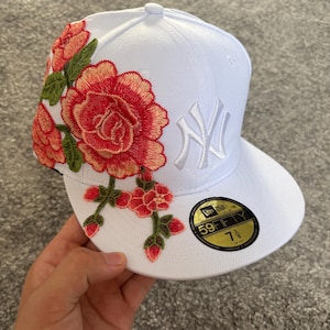 Könnte beinhalten: Eine weiße New Era 59FIFTY Cap mit einem gestickten New York Yankees Logo in Weiß und einem großen Blumenmuster in Pink, Orange und Grün an der Seite. Ein schwarz-goldener Aufkleber auf dem Schirm zeigt die Größe 58,7 cm an.