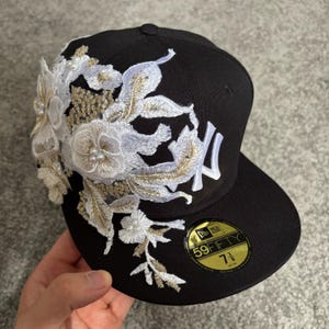 Peut inclure: Une casquette de baseball noire ornée d'une broderie florale blanche et dorée sur le côté gauche, agrémentée de petites perles blanches. La casquette présente un logo New York Yankees blanc et un autocollant 59FIFTY indiquant une taille de 58,7 cm.