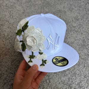 May include: A white baseball cap featuring an embroidered New York Yankees logo on the front and large white floral appliques with green leaves on the side. The flat brim includes a circular black and gold 59FIFTY sticker with the size 7 1/8.