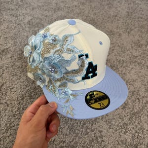 Op de afbeelding: Een crèmekleurige honkbalpet met een lichtblauwe klep en een lichtblauwe knoop aan de bovenkant. De pet is voorzien van ingewikkeld lichtblauw bloemenborduurwerk met gouden accenten en kleine parels. De letters LA zijn in zwart en blauw geborduurd. Een 59FIFTY 7 1/8 sticker zit op de klep.