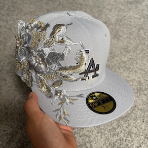 Könnte beinhalten: Eine hellgraue Baseballkappe mit einer floralen Stickerei in Silber- und Goldfäden sowie weißen Perlenakzenten. Die Vorderseite zeigt das LA-Logo in Dunkelbraun und Weiß, und ein schwarz-goldener 59FIFTY-Aufkleber gibt die Größe 57,7 cm an.