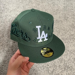 May include: A forest green fitted baseball cap featuring a white embroidered LA logo on the front, dark green script text on the side, and a black and gold 59FIFTY sticker on the brim indicating a size of 7 3/8 inches.