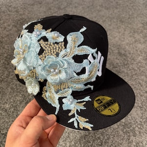 Puede incluir: Una gorra de béisbol negra con un diseño floral bordado en hilo azul claro y dorado con detalles de perlas blancas. La gorra muestra un logotipo blanco de los New York Yankees y una pegatina circular 59FIFTY en la visera que indica la talla 7 1/2.