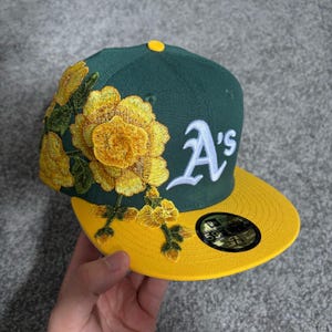 Pode incluir: Um boné de beisebol verde-escuro com aba amarela e um logótipo A's bordado a branco. O lado esquerdo do boné está decorado com um grande e detalhado bordado floral amarelo. Um autocolante circular preto e dourado está na aba plana.