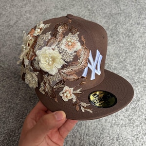 Peut inclure: Une casquette de baseball marron avec un logo New York Yankees brodé en blanc sur le devant. Le côté gauche est orné d'une broderie florale complexe dans des tons crème et marron, agrémentée de petites perles blanches sur tout le motif.
