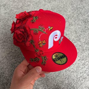 Op de afbeelding: Een felrode honkbalpet met een grote witte geborduurde letter P op de voorkant. De pet is versierd met gedetailleerd borduurwerk van rode rozen, groene stengels en een kleine bij. Een zwarte 59FIFTY-sticker met maat 7 1/4 is op de klep bevestigd.