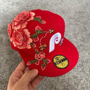 Op de afbeelding: Een felrode honkbalpet met een grote witte geborduurde letter P, een kleine bij en gedetailleerd bloemenborduurwerk in roze en oranje tinten. Een ronde zwart-gouden sticker op de platte klep toont de tekst 59FIFTY 7 3/8 58.7cm.
