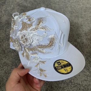 Può includere: Un cappellino da baseball bianco con un motivo floreale ricamato sul lato sinistro, arricchito da fili dorati e piccole perle bianche. Il cappellino presenta il logo dei New York Yankees sul davanti e un adesivo circolare nero e oro 59FIFTY.