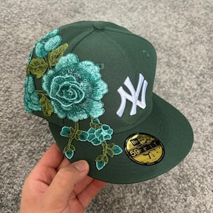 Pode incluir: Um boné de beisebol verde-escuro com o logótipo dos New York Yankees bordado a branco na frente e um grande bordado floral azul-petróleo na lateral. Um autocolante circular preto e dourado 59FIFTY de tamanho 7 1/4 está na aba.