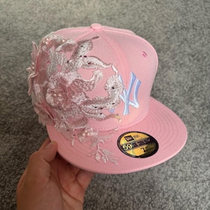 Op de afbeelding: Een lichtroze New Era 59FIFTY honkbalpet met een wit geborduurd New York Yankees logo aan de voorzijde en een gedetailleerde lichtroze kanten bloemenapplicatie versierd met kleine witte parels aan de linkerkant van de kroon en de klep.