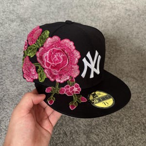 Op de afbeelding: Een zwarte New Era 59FIFTY honkbalpet met een wit geborduurd New York Yankees logo op de voorkant en levendige roze rozenborduurwerken met groene stelen en bladeren die vanaf de zijkant doorlopen op de platte klep, voorzien van een 7 3/8 sticker.