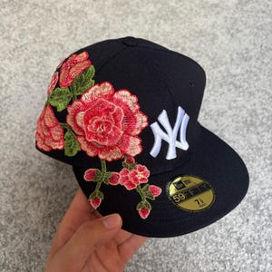 Pode incluir: Um boné de beisebol preto com o logótipo dos New York Yankees bordado a branco e um grande padrão floral com rosas cor-de-rosa e folhas verdes. Um autocolante dourado e preto na pala exibe o texto 59FIFTY e o tamanho 7 1/2.