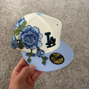 Op de afbeelding: Een crèmekleurige honkbalpet met een lichtblauwe klep en een opvallend zwart en lichtblauw LA-logo. De zijkant is voorzien van gedetailleerd blauw bloemenborduurwerk met groene bladeren. Een zwarte 59FIFTY-sticker op de klep toont maat 57,7 cm.