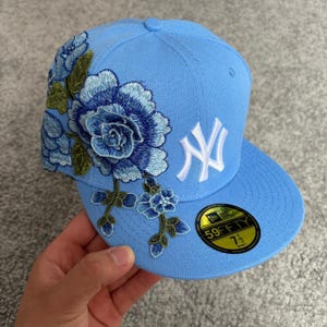Pode incluir: Um boné de beisebol azul claro com um design floral bordado em tons de azul e verde na lateral. A frente exibe um logotipo branco dos New York Yankees. Um adesivo circular preto e dourado 59FIFTY na aba mostra 59,6 cm.