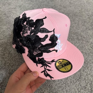 Può includere: Un cappellino da baseball rosa chiaro con una vistosa applicazione in pizzo floreale nero sul lato. Il cappellino presenta un logo ricamato bianco e un adesivo circolare oro e nero sulla visiera piatta che indica la taglia 7 3/8, 58,7 cm.