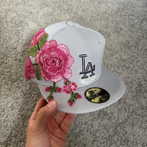 Op de afbeelding: Een lichtgrijze honkbalpet met een geborduurd Los Angeles Dodgers-logo in wit met een zwarte omlijning aan de voorzijde. De linkerkant is versierd met felroze rozenborduursels met groene bladeren en stengels. Een zwarte sticker zit op de klep.