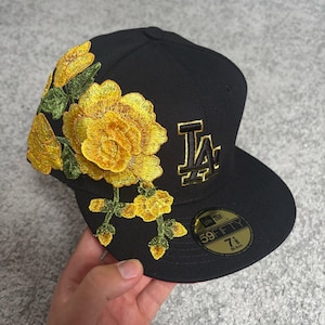 Può includere: Un cappellino da baseball nero con un motivo floreale giallo ricamato sul lato e il logo LA con contorno dorato sul davanti. Un adesivo circolare sulla visiera piatta riporta il testo 59FIFTY, 7 1/8 e 56,8 cm, indicando la taglia specifica.