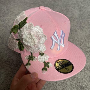 Op de afbeelding: Een lichtroze New Era 59FIFTY honkbalpet met een geborduurd wit bloemmotief en groene bladeren aan de zijkant. De voorkant toont een wit NY-logo en een ronde zwart-gouden sticker op de klep geeft de maat 57,7 cm aan.