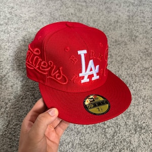 May include: A bright red fitted baseball cap featuring a white embroidered LA logo on the front. The side displays the word Dodgers in red script embroidery. A black and gold 59FIFTY sticker is on the flat brim, indicating a size of 7 1/4.
