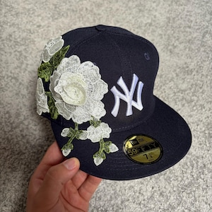 Może przedstawiać: Granatowa czapka z daszkiem z białym haftowanym logo New York Yankees z przodu oraz dużą białą aplikacją kwiatową z zielonymi łodygami z boku. Okrągła złoto-czarna naklejka na płaskim daszku zawiera napisy 59FIFTY oraz 7 1/8.