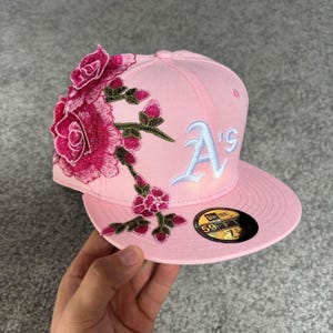Peut inclure: Une casquette de baseball rose clair avec un logo A's bleu clair brodé sur le devant et un grand motif floral composé de roses rose foncé et de tiges vertes sur le côté gauche. Un autocollant 59FIFTY noir et or est sur la visière plate.