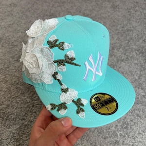 Può includere: Un cappellino da baseball turchese brillante con il logo dei New York Yankees ricamato in bianco e una grande e dettagliata applicazione floreale bianca con steli e boccioli verdi sul lato. Un adesivo nero e oro sulla visiera indica la taglia 7 1/4.
