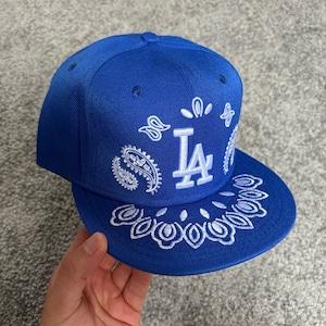 Pode incluir: Um boné de beisebol azul royal com um logótipo LA bordado a branco no painel frontal. O boné é decorado com um padrão paisley branco que se estende dos painéis frontais até à aba plana, criando um design detalhado e muito coeso.
