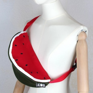 May include: A novelty fanny pack shaped like a watermelon, with a red body, white rind, and black seed details. The bag is olive green on the bottom and has a red strap. The brand name "ACORN" is printed on the bag.