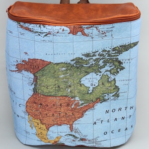 Puede incluir: Mochila rectangular con un mapa de América del Norte sobre un fondo azul claro. La parte superior y el asa son marrones. El mapa incluye Estados Unidos, Canadá y los océanos circundantes. Cierre con cremallera.