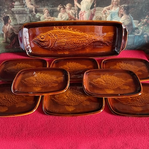 Peut inclure: Un ensemble de plats de service en céramique marron. L'ensemble comprend un grand plat rectangulaire et sept petites assiettes rectangulaires, tous avec un motif de poisson détaillé. Les plats sont disposés sur une surface rouge.