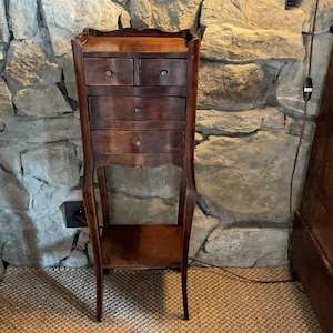 May include: A tall, dark brown wooden chest of drawers featuring two small top drawers, two larger drawers below, and a lower open shelf. The piece stands on four slender, curved legs and includes a flat top surface with a raised edge border.