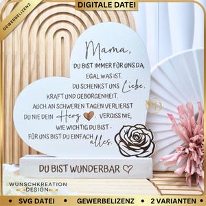 Puede incluir: Placa de madera en forma de corazón con texto en alemán, apoyada sobre una base rectangular blanca. La placa presenta un diseño de rosa y las palabras "Mama, Du bist wunderbar". El fondo incluye un arco de madera y elementos decorativos.