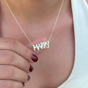 Op de afbeelding: Een goudkleurige ketting met de naam "MARRY" in een gestileerd lettertype. De hanger hangt aan een fijne ketting. De persoon draagt een witte top.
