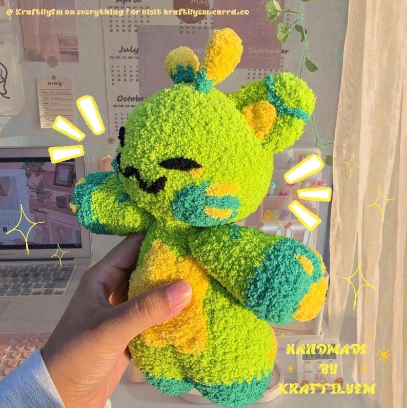 May include: A handmade plush toy in the shape of a bear, featuring a vibrant color scheme of green, yellow, and teal. The bear has a smiling face with black embroidered details. The text "HANDMADE BY KRAFTILYSM" is visible on the bottom right.
