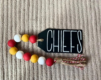 May include: A decorative wooden bead garland with a black tag that reads "CHIEFS" in white lettering. The garland features red, yellow, and white beads. A tassel with red and white twine accents the end of the garland.