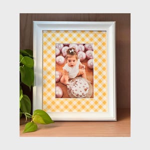 Può includere: Foto con cornice bianca con un bambino e una torta, circondata da palloncini rosa. La cornice ha un motivo a scacchi giallo e bianco. La foto è esposta su una superficie di legno, con una pianta verde nell'angolo.