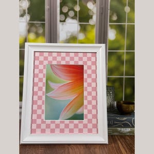Può includere: Un quadro con cornice bianca con un passe-partout rosa a scacchi. L'immagine all'interno della cornice è un primo piano di un fiore con petali arancioni, gialli e verdi. La cornice è su una superficie di legno.