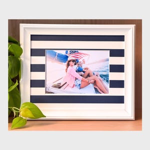 Può includere: Foto con cornice bianca con un passe-partout a righe blu navy e bianche. La foto mostra persone su una barca con una bandiera americana. La cornice è su una superficie di legno con foglie verdi.