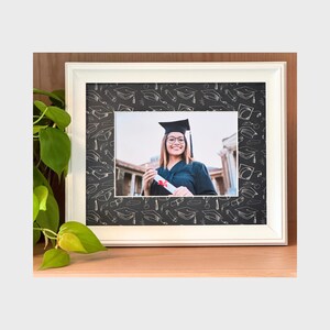 Può includere: Foto con cornice bianca di una laureata con cappello e toga, che tiene in mano un diploma. La cornice ha uno sfondo nero con illustrazioni bianche di cappelli di laurea. La foto è su una superficie di legno con foglie verdi.