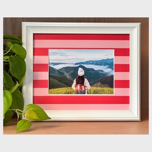 Può includere: Un quadro con cornice bianca con una fotografia di una persona che guarda una catena montuosa. La cornice ha un bordo a righe rosse e rosa. La fotografia è di una persona con un cappello bianco e uno zaino. La cornice è su una superficie di legno.