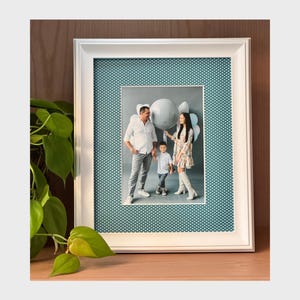 Può includere: Un ritratto di famiglia incorniciato. La foto mostra una famiglia di tre persone in posa con palloncini. La foto è ambientata su uno sfondo verde acqua con pois bianchi, incorniciata in un bordo bianco. La cornice è esposta su una superficie di legno.