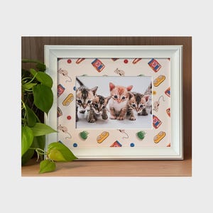 Può includere: Un'immagine con cornice bianca con quattro gattini. La cornice ha un bordo a motivi con illustrazioni di cibo per gatti, topi e altri elementi relativi ai gatti. I gattini sono un mix di colori arancione e tabby.