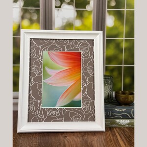 Può includere: Un quadro con cornice bianca con stampa floreale. L'immagine presenta un primo piano di un fiore con petali arancioni, gialli e verdi. La cornice ha uno sfondo marrone con un motivo a rose bianche.